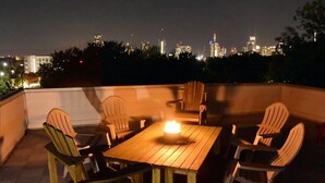 Une terrasse sur le toit avec une table et des chaises en bois, un petit foyer et, en arrière-plan, la vue sur les toits de la ville.