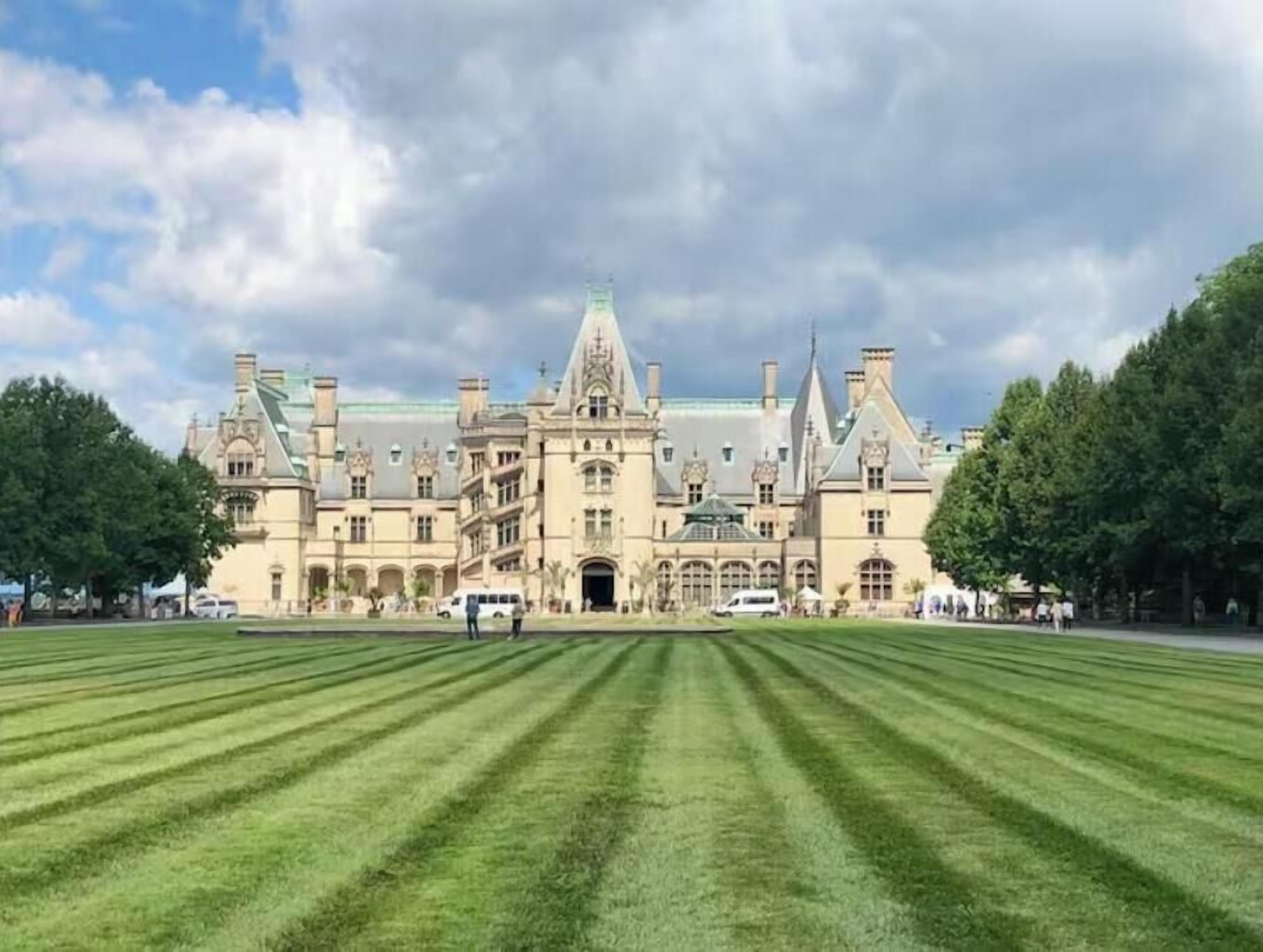 A large, historic building with a symmetrical facade, multiple towers, and a well-maintained lawn in front.