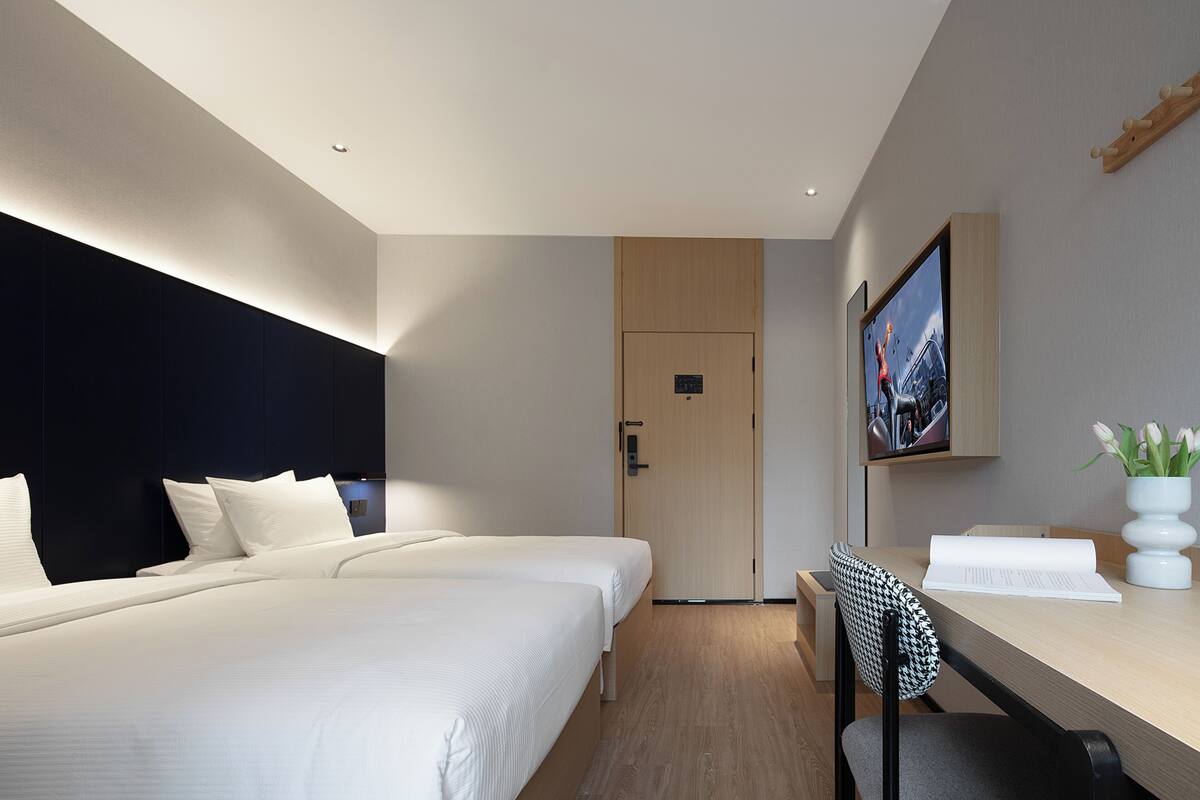 A modern hotel room with two beds, a desk, a chair, a television, and a door.