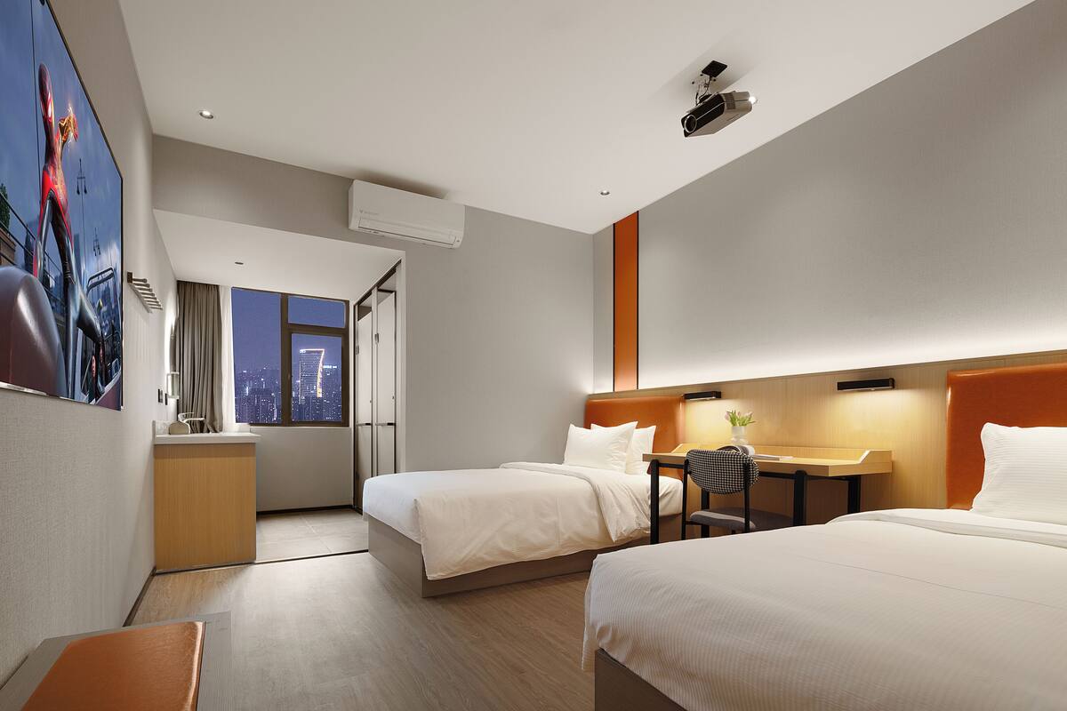 A hotel room with two beds, a desk, a chair, and a view of the cityscape.