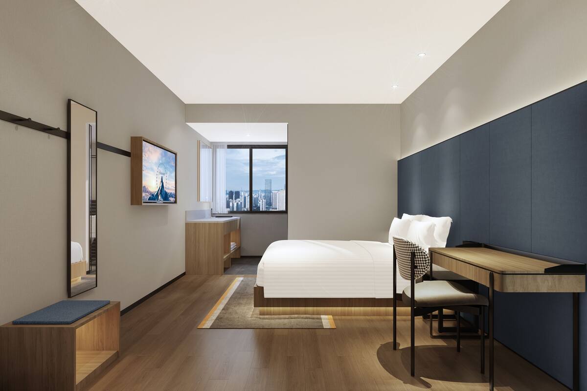 A modern hotel room with a bed, desk, chair, and city view through the window.