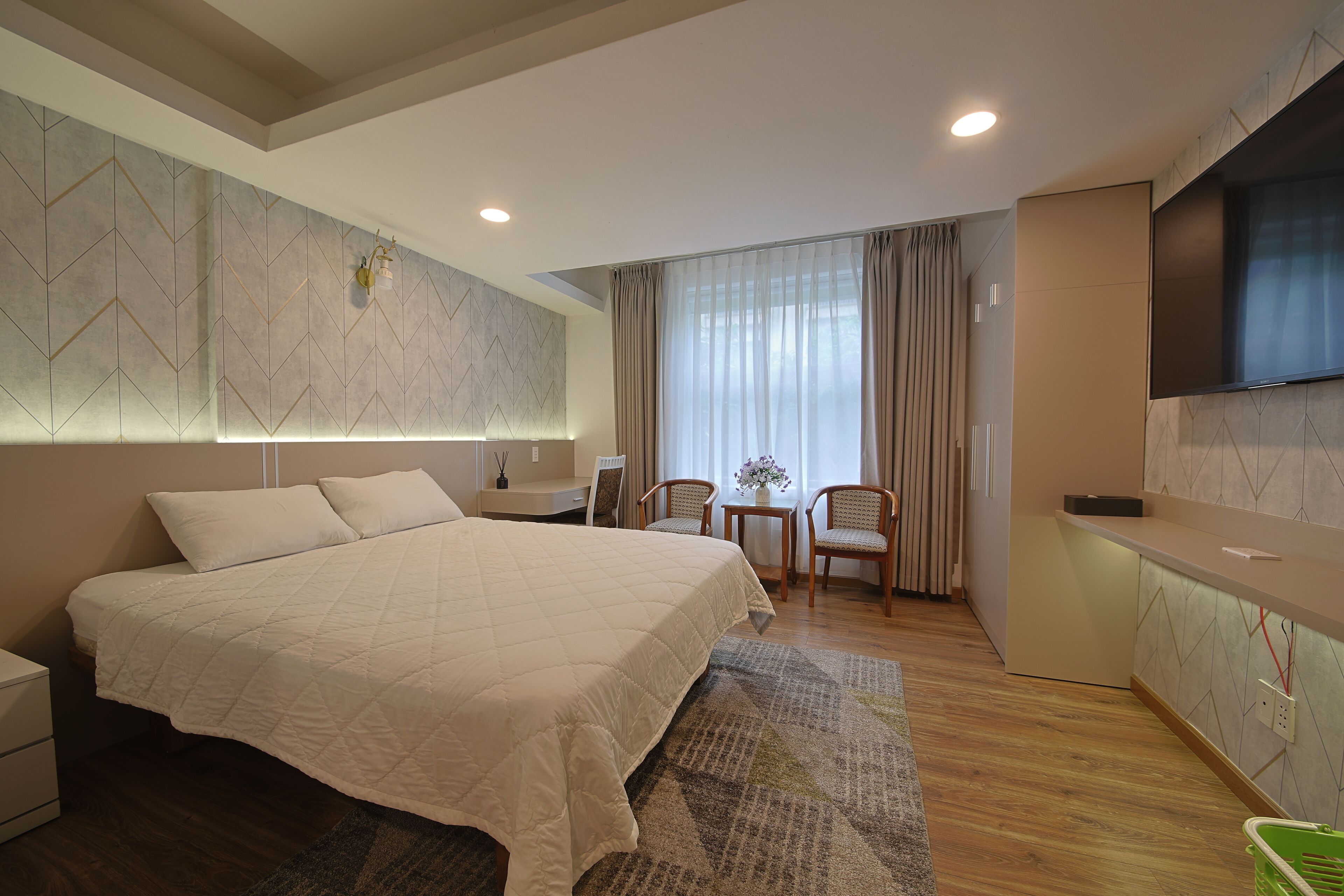 A modern hotel room with a large bed, a desk, a chair, a small table with a vase, and a TV mounted on the wall.