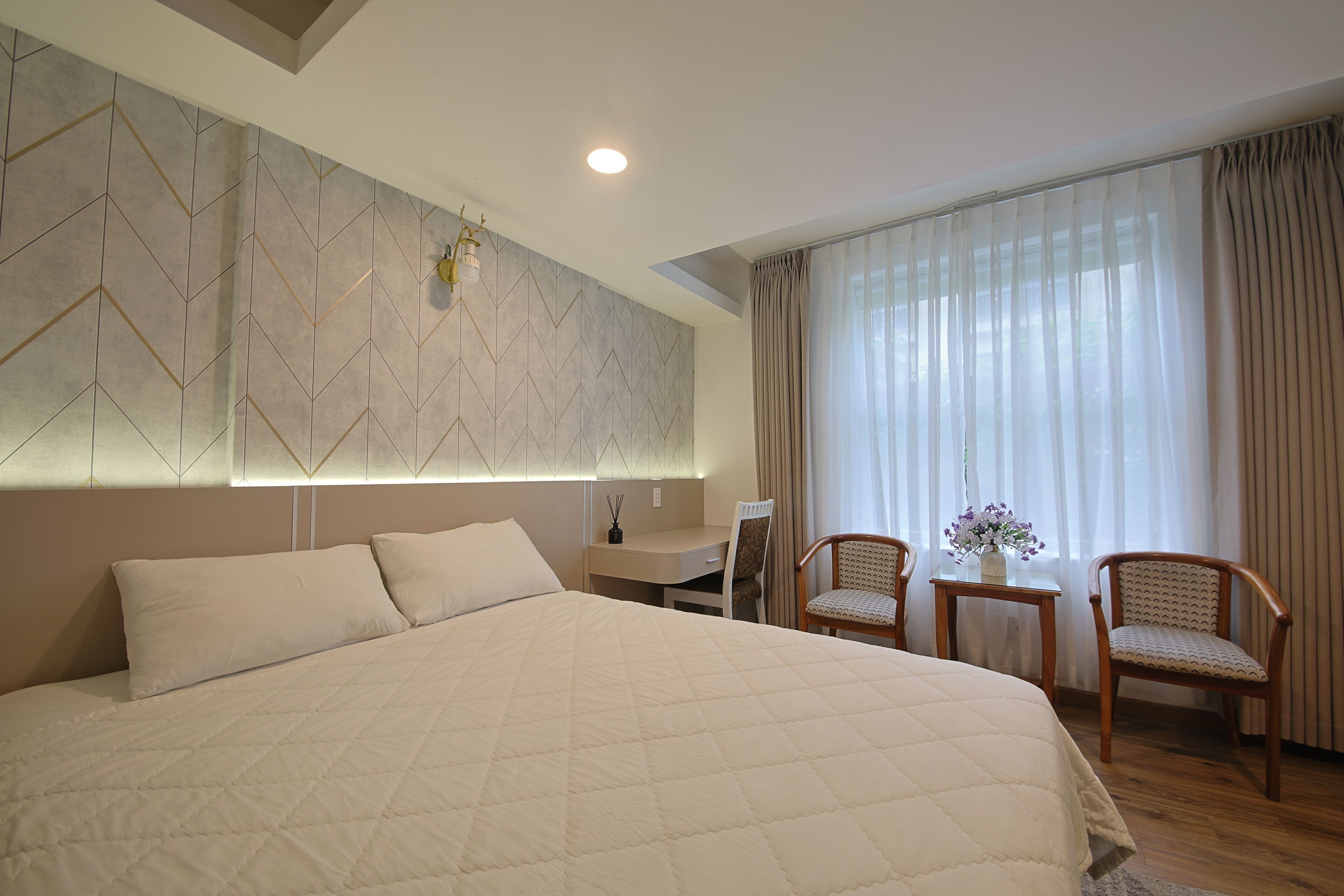 Habitación de hotel moderna con una cama grande, un escritorio, dos sillas y vistas al exterior a través de cortinas translúcidas.