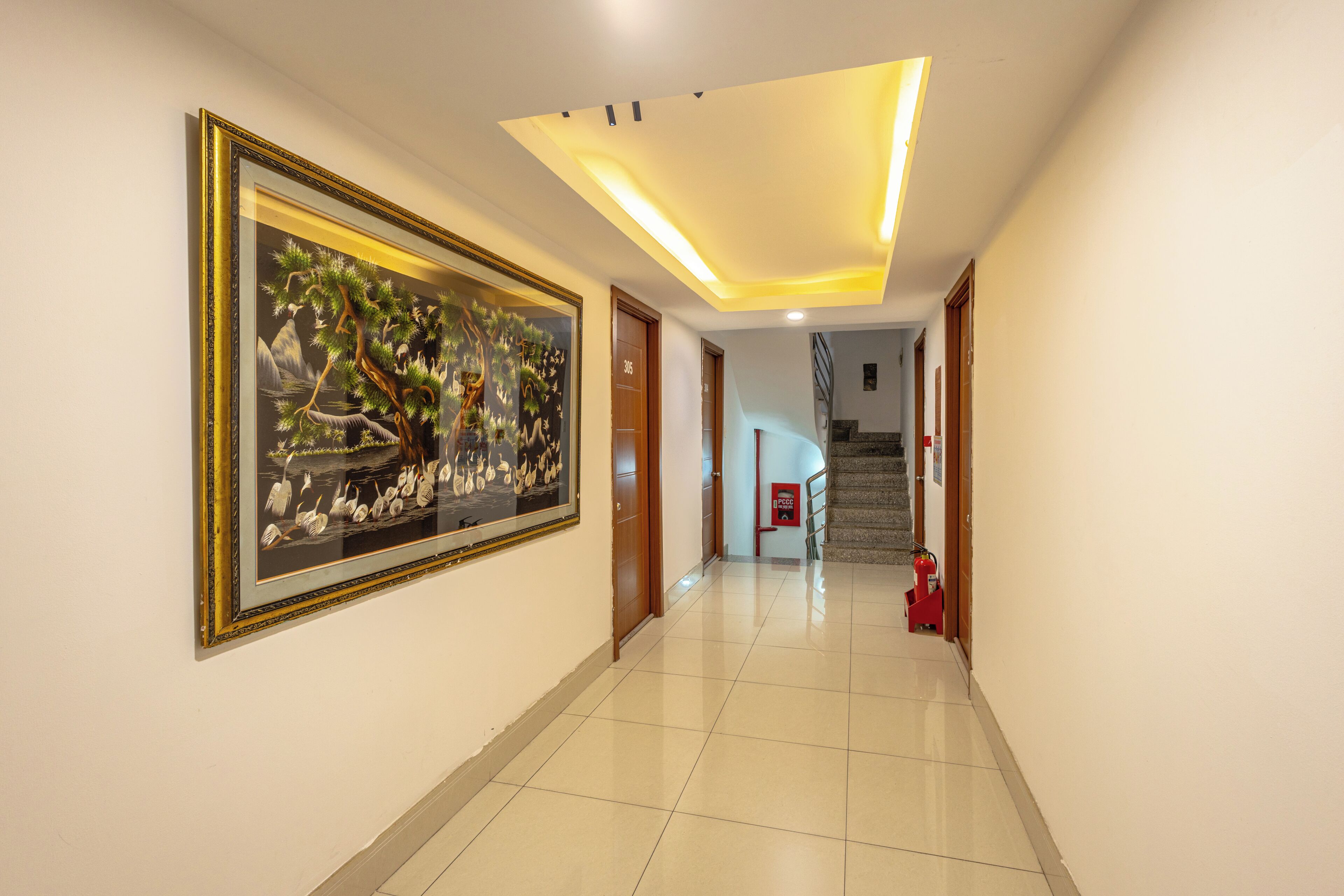 A hotel corridor with a framed artwork on the wall, a fire extinguisher, and a staircase in the background.