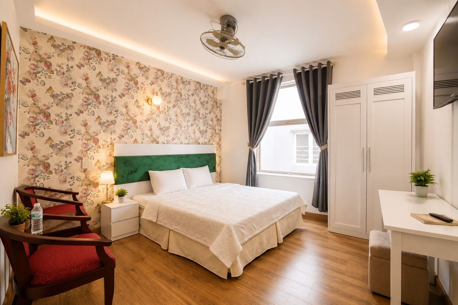 A hotel room with a bed, a desk, a chair, a nightstand, a lamp, a potted plant, and a window with curtains.