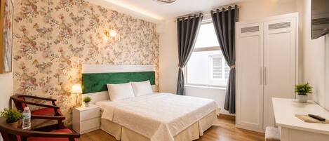 Ein Hotelzimmer mit Bett, Schreibtisch, Stuhl, Nachttisch, Lampe, einer Zimmerpflanze und einem Fenster mit Vorhängen.