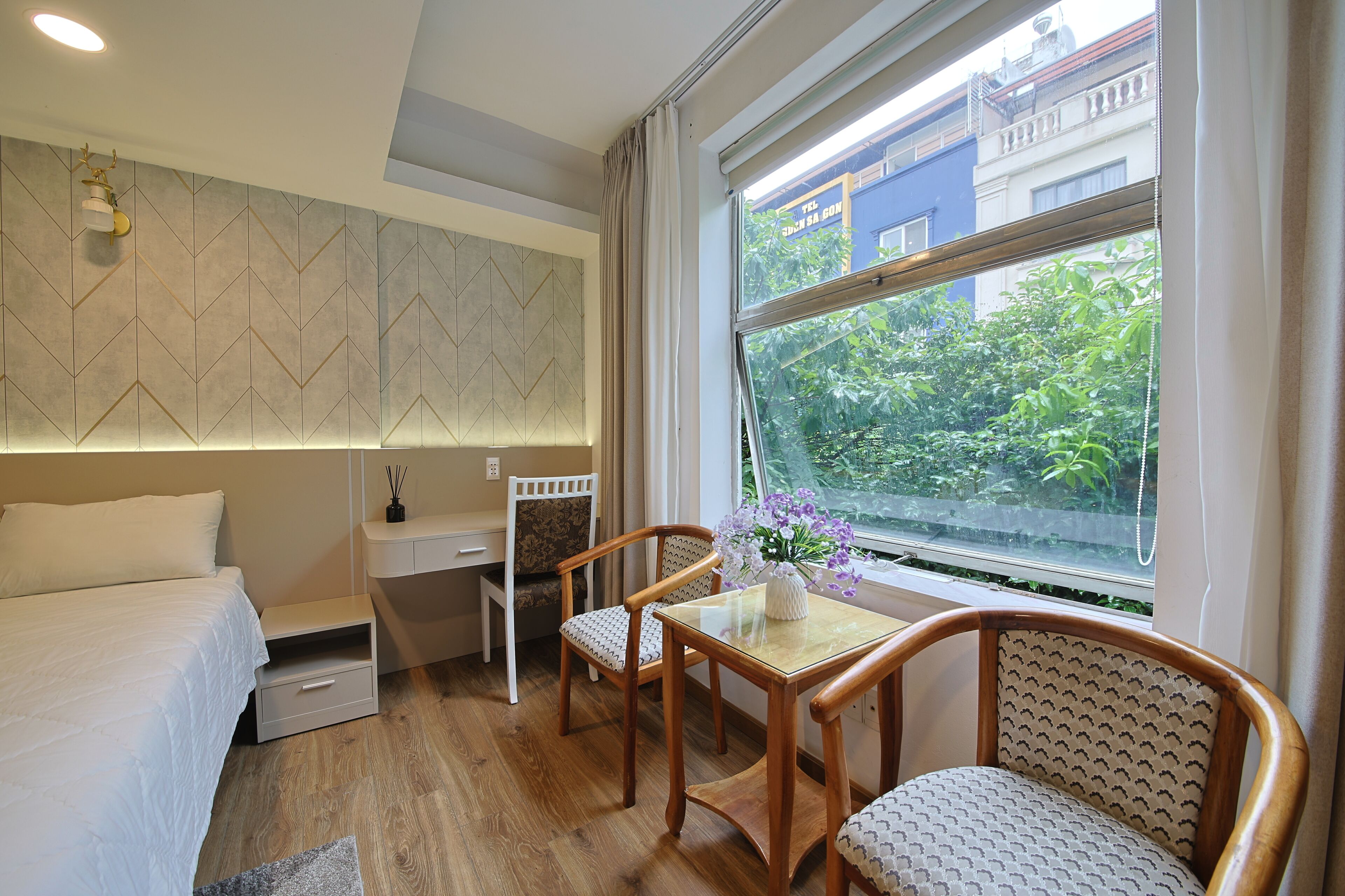 A bedroom with a bed, desk, two chairs, a small table, and a window with a view of greenery and buildings.