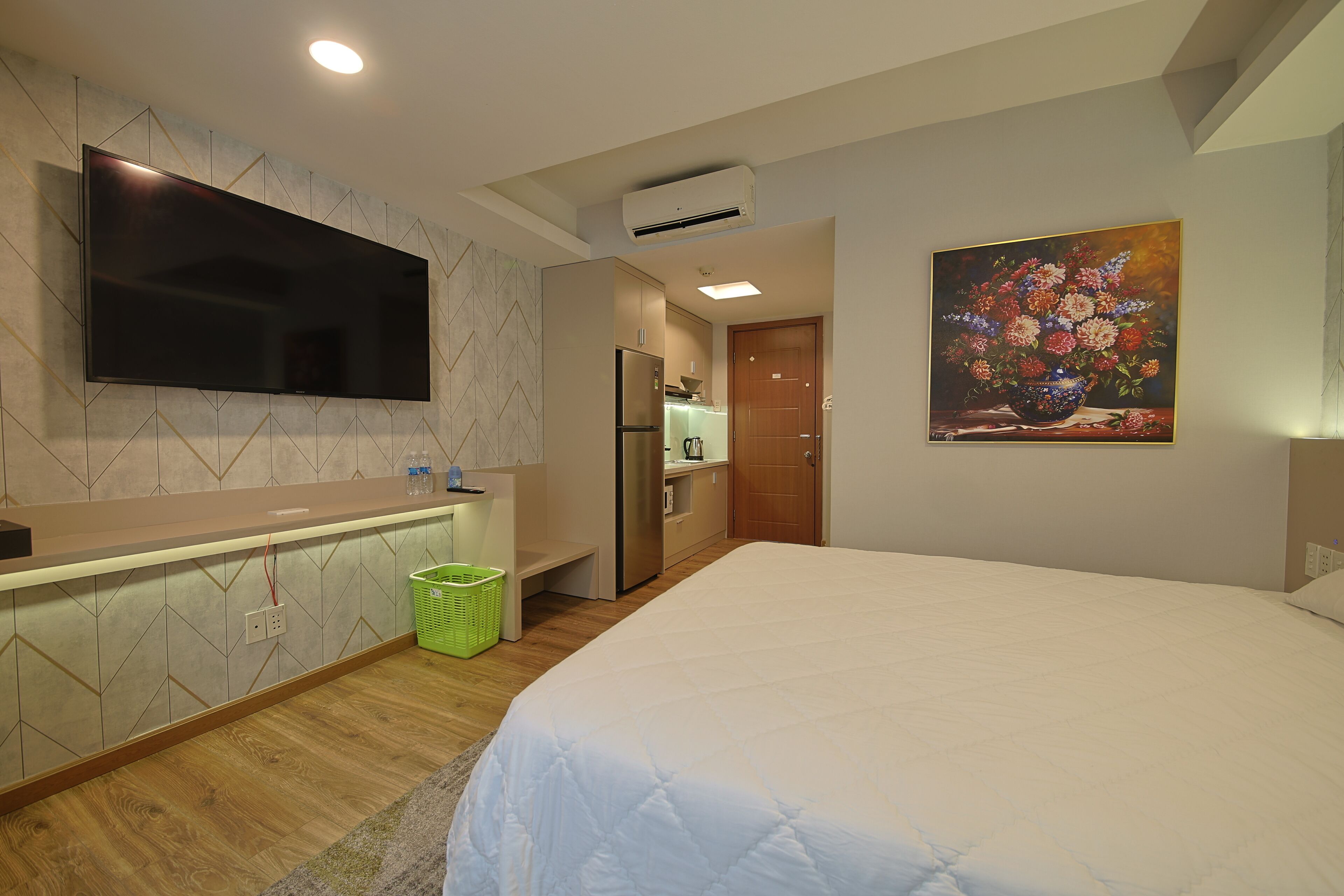 Habitación de hotel moderna con un televisor de pantalla plana grande montado en la pared, una cama con ropa blanca, una kitchenette pequeña y un cuadro de flores en la pared.
