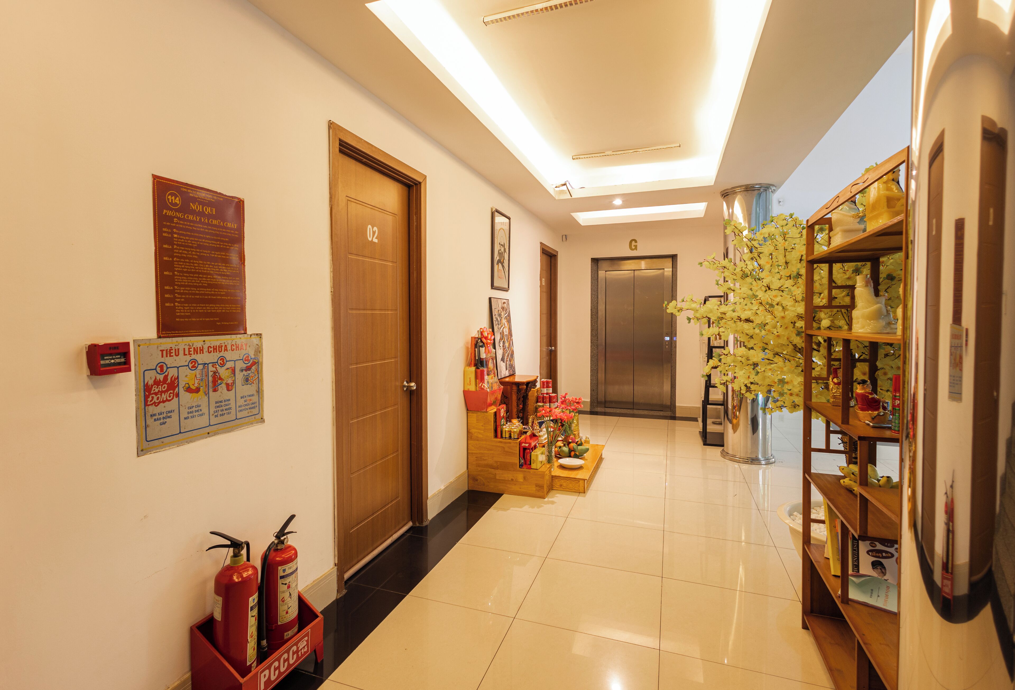 A hotel corridor with a wooden door labeled "02", a fire extinguisher, a wooden cabinet with flowers, and a bookshelf with various items.