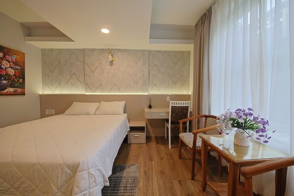 A neatly arranged hotel room with a large bed, a desk with a chair, a vase of flowers, and a window with curtains.