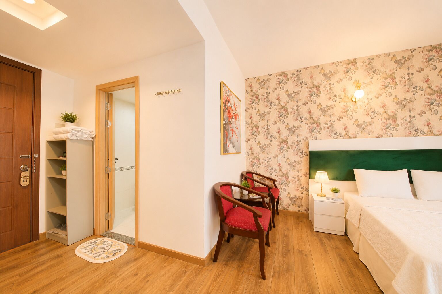 Hotelkamer met houten vloer, een bed met witte lakens, nachtkastje met lamp, stoel met rode kussens, houten deur, en plank met handdoeken en plant.
