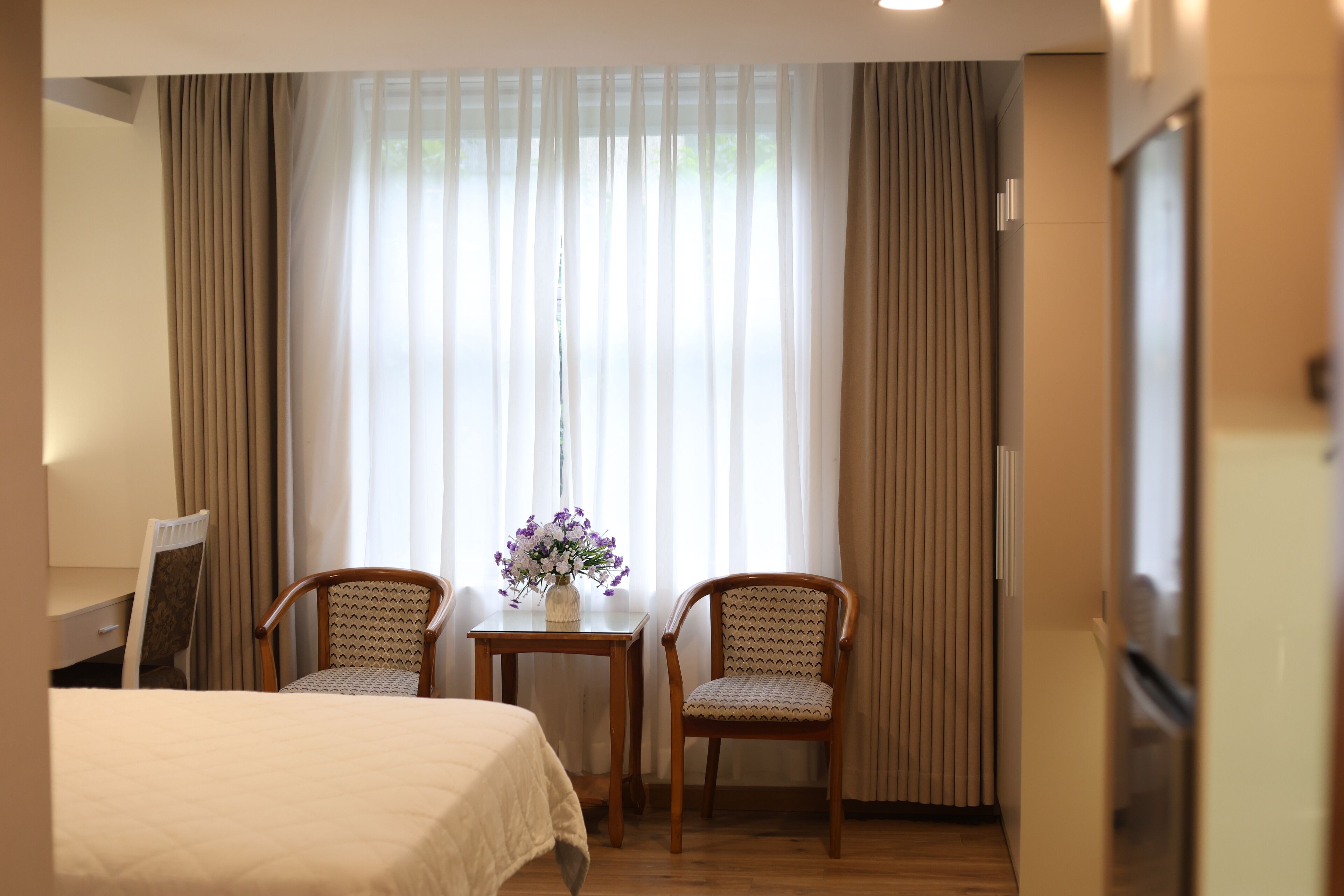 A hotel room with a bed, two chairs, a small table with a vase of flowers, and a window with curtains.