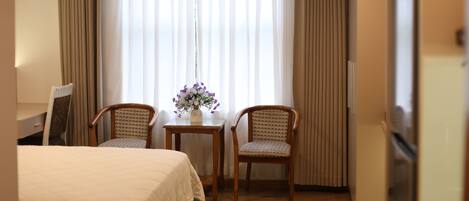 A hotel room with a bed, two chairs, a small table with a vase of flowers, and a window with curtains.