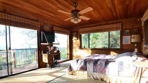 A wooden cabin bedroom with a bed, a ceiling fan, a window with a view, and a small table with a lamp. - Charming Rustic Mountain Cabin, Near Yosemite,Amazing Views, and a Fireplace (Wishon)
