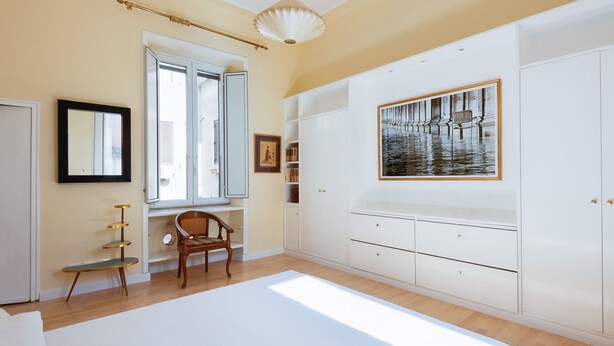 A bedroom with a large bed, a window with blinds, a chair, a white wardrobe, and a framed picture on the wall. - Babuino by Interhome (Roma: Piazza di Spagna)