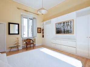 Un dormitorio con una cama grande, una ventana con persianas, una silla, un armario blanco y un cuadro en la pared.