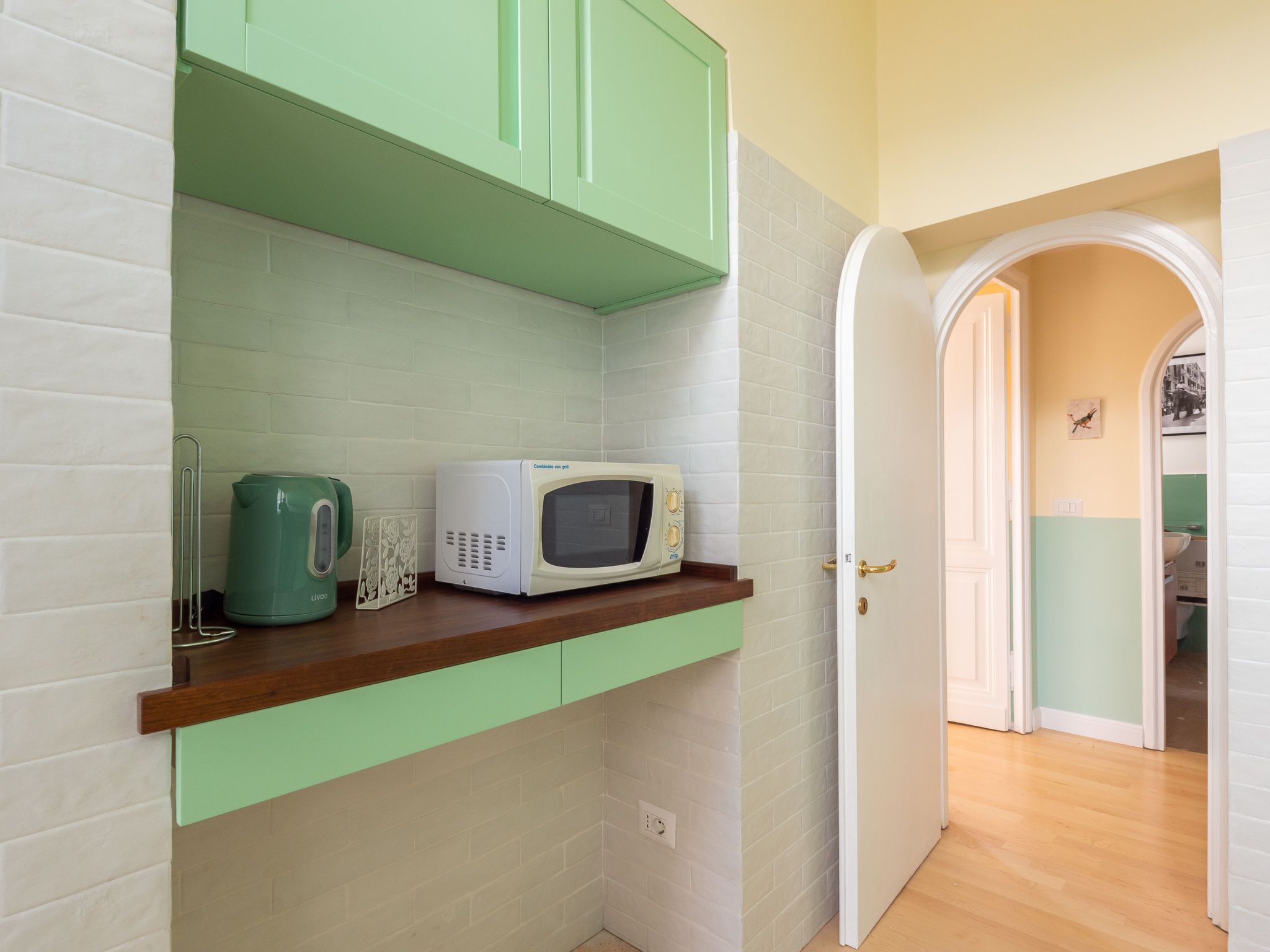 A kitchen with a microwave on a shelf, a kettle, and a cabinet.