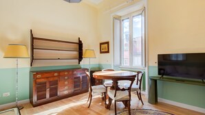 A dining area with a wooden table, chairs, a cabinet with drawers, a television, and a window with a view of buildings. - Babuino by Interhome (Roma: Piazza di Spagna)