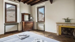 A room with a wooden ceiling, a fireplace, a large window with a view of a building, a dresser with a mirror, and a bed with striped bedding. - Pantheon View by Interhome (Roma: Piazza Navona - Campo dei Fiori)