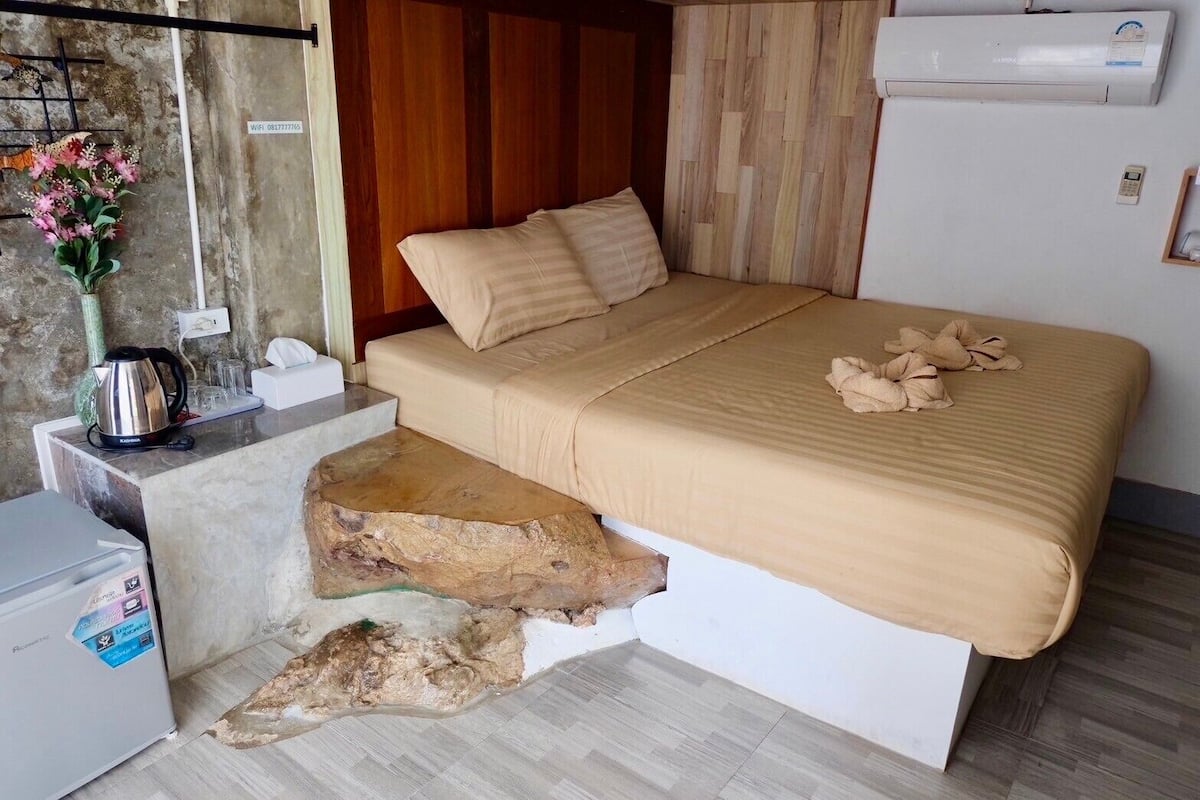 A bedroom with a bed, pillows, a kettle, a small fridge, and a decorative vase.