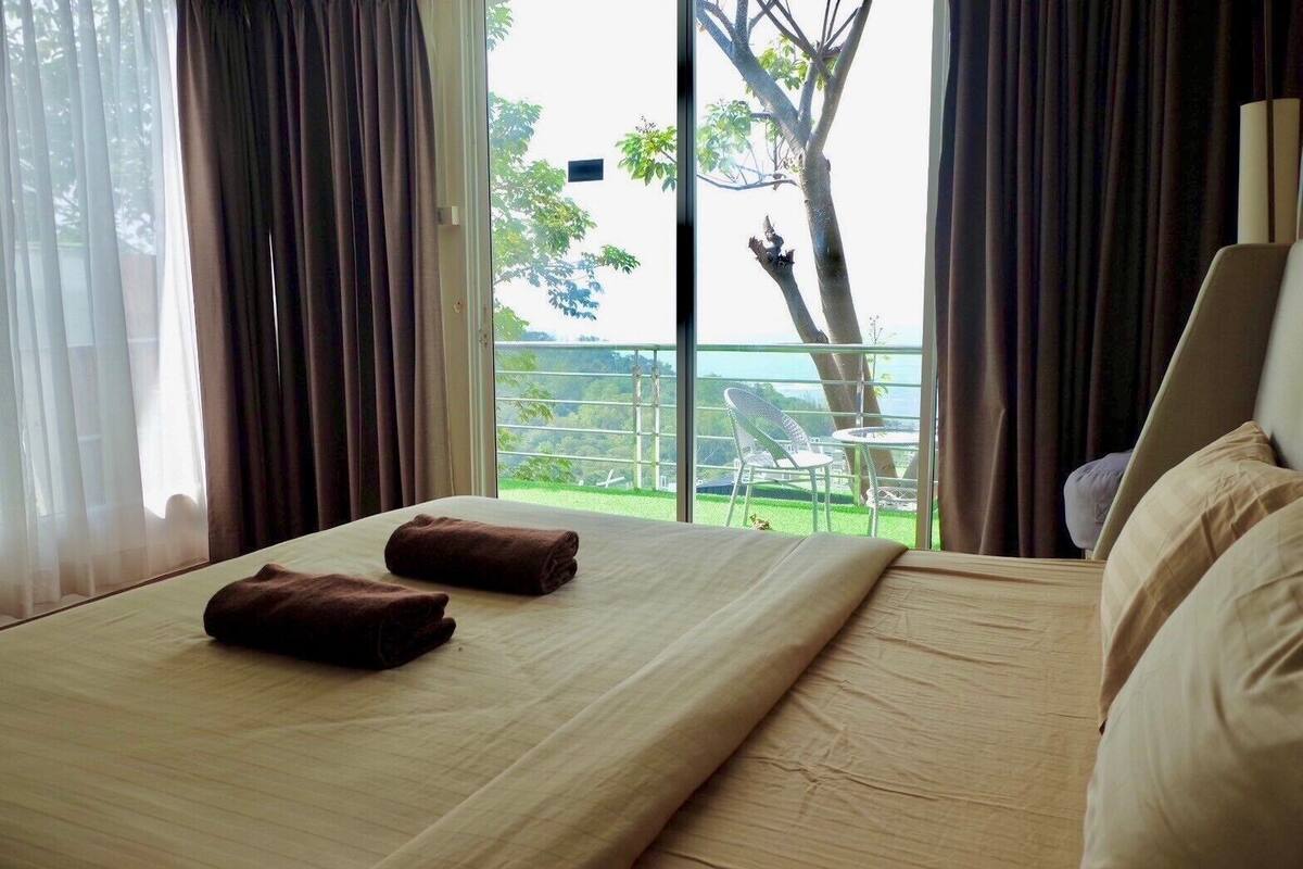 A hotel room with a bed, two towels, a window with curtains, and a view of a balcony with outdoor furniture.