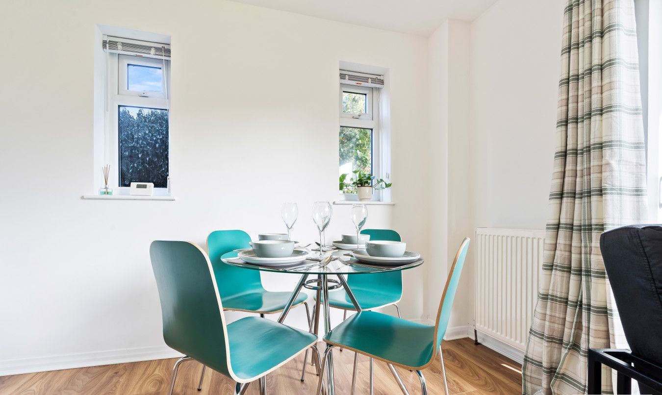 A dining area with a round table, teal chairs, and a window with a view of trees.