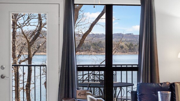 A room with a view of a lake and hills, a table with chairs, and a sofa. - Lakefront Condo, 2 min to Silver Dollar City! Ticket Deals & Resort Perks (Branson)