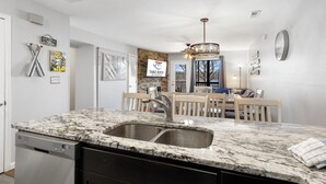A kitchen with a granite countertop, stainless steel sink, and a dishwasher. There is a dining area with a table and chairs, and a sign on the wall that reads "Table Rock Resort." - Lakefront Condo, 2 min to Silver Dollar City! Ticket Deals & Resort Perks (Branson)