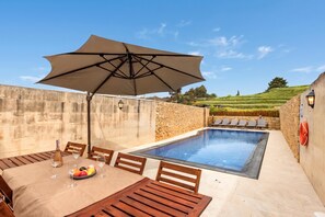 Un espace aménagé au bord de la piscine, avec une table dressée pour les repas, un grand parasol et un mur en pierre.