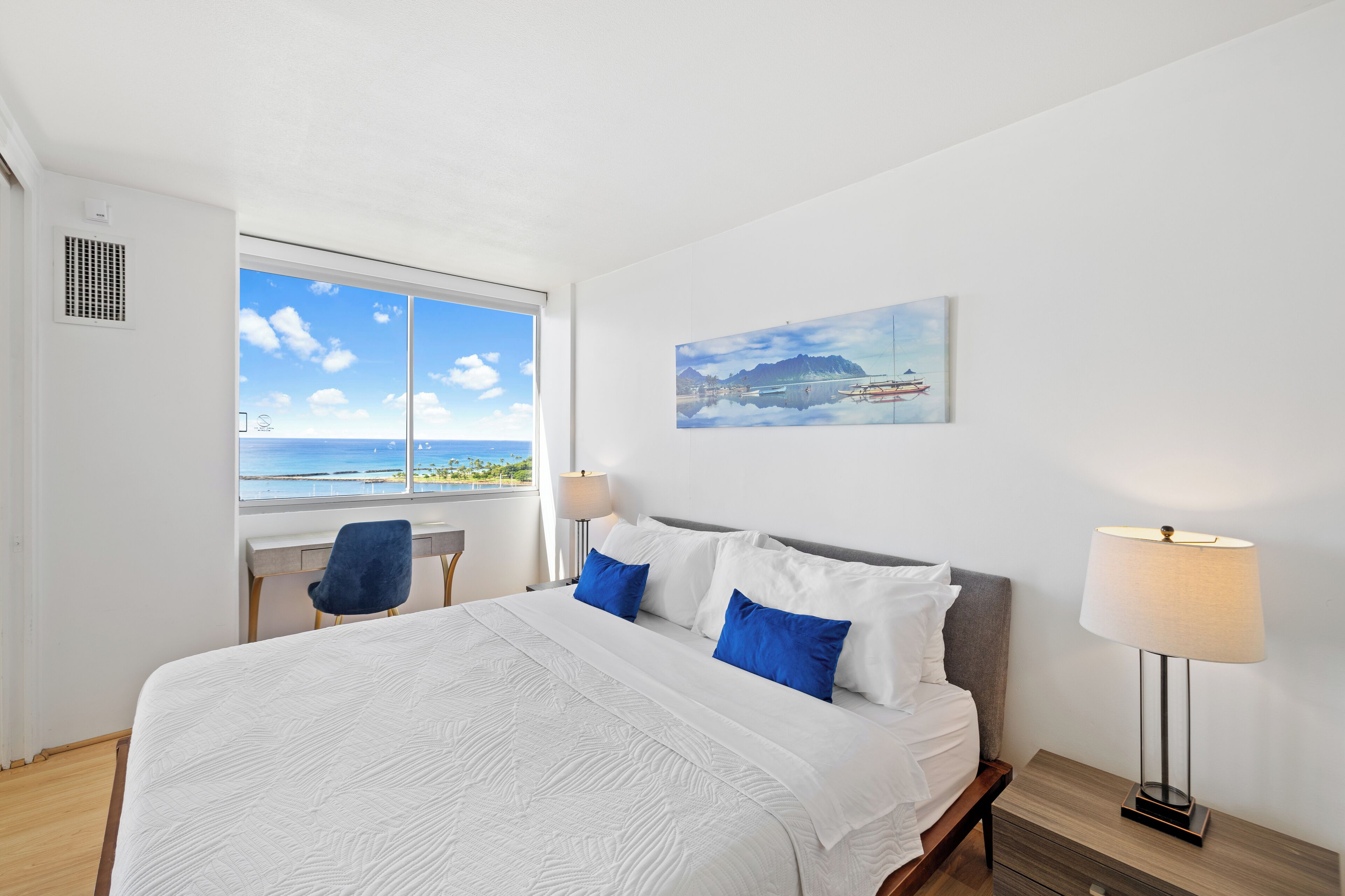 A hotel room with a large bed, a desk, and a chair. There is a window with a view of the ocean and a painting on the wall.