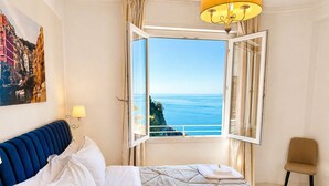 A bedroom with a bed, a chair, a window with an open view, a painting, and a lamp. - Vertigini Suites (Riomaggiore)