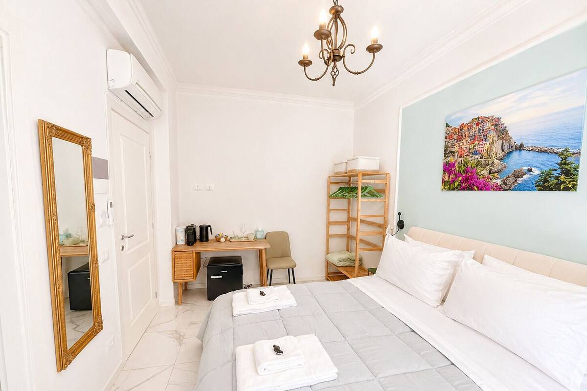 Ein Hotelzimmer mit Bett, Schreibtisch, Stuhl, Kleiderschrank, Spiegel, einer Chandelierleuchtre und einem Gemälde mit einer Küstenansicht.