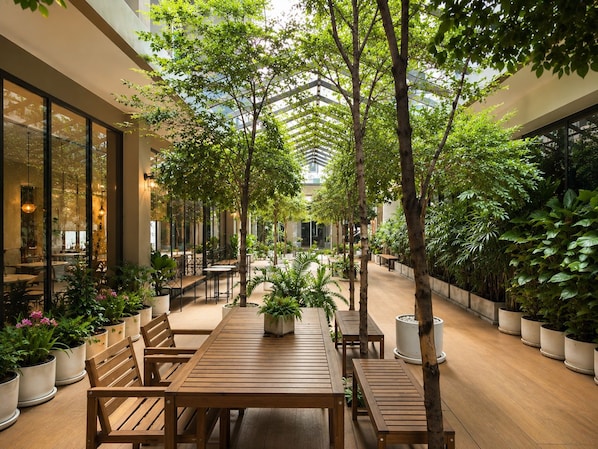 Un jardín interior con mesas y sillas de madera, rodeado de plantas y árboles en macetas.