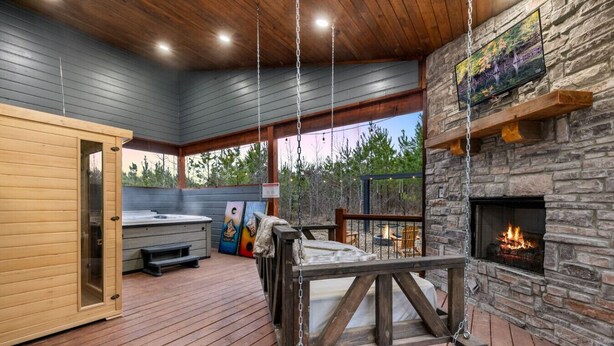 A wooden deck with a hot tub, a stone fireplace, and a swing chair. - Over the Moon · Sauna & Hot Tub at Modern Intimate Escape (Broken Bow)