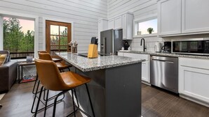 A modern kitchen with white cabinetry, a granite countertop, and stainless steel appliances. There are bar stools and a large window providing a view of the outdoors. - Over the Moon · Sauna & Hot Tub at Modern Intimate Escape (Broken Bow)