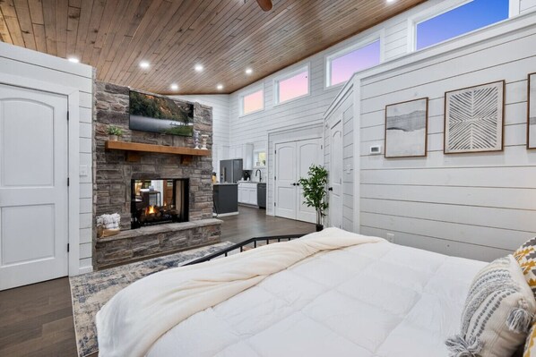 A bedroom with a large bed, a fireplace, a kitchen area, and a view of the outdoors.