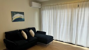 Ein Wohnzimmer mit schwarzem Sofa, beigen Kissen, einem Gemälde an der Wand und einem großen Fenster mit weißen Vorhängen.