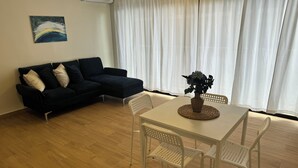 Ein Wohnzimmer mit einem dunkelgrauen Sofa, einem weißen Esstisch mit Stühlen, einer Vase mit Blumen und einem Bild an der Wand.