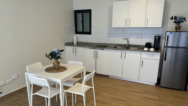 Eine kleine Küche mit weißen Schränken, einem Edelstahlkühlschrank, einer Granit-Arbeitsplatte und einem Essbereich mit weißen Stühlen und einem Tisch.