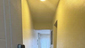 Ein schmaler Korridor mit Holzdielen führt zu einem Zimmer mit einem Bett und einem Fenster mit Vorhängen.