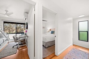 A modern bedroom with a bed, a desk, and a chair. There is a window with a view of trees. - SUMMER SALE! 3BR Condo w Sweeping Ocean Views Near the Beach - Kaanapali Plantation 32 (Lahaina)