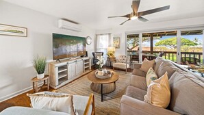 A spacious living room with a large sofa, a coffee table, and a television. There is a ceiling fan, a plant, and a view of the outdoors through large windows. - SUMMER SALE! 3BR Condo w Sweeping Ocean Views Near the Beach - Kaanapali Plantation 32 (Lahaina)