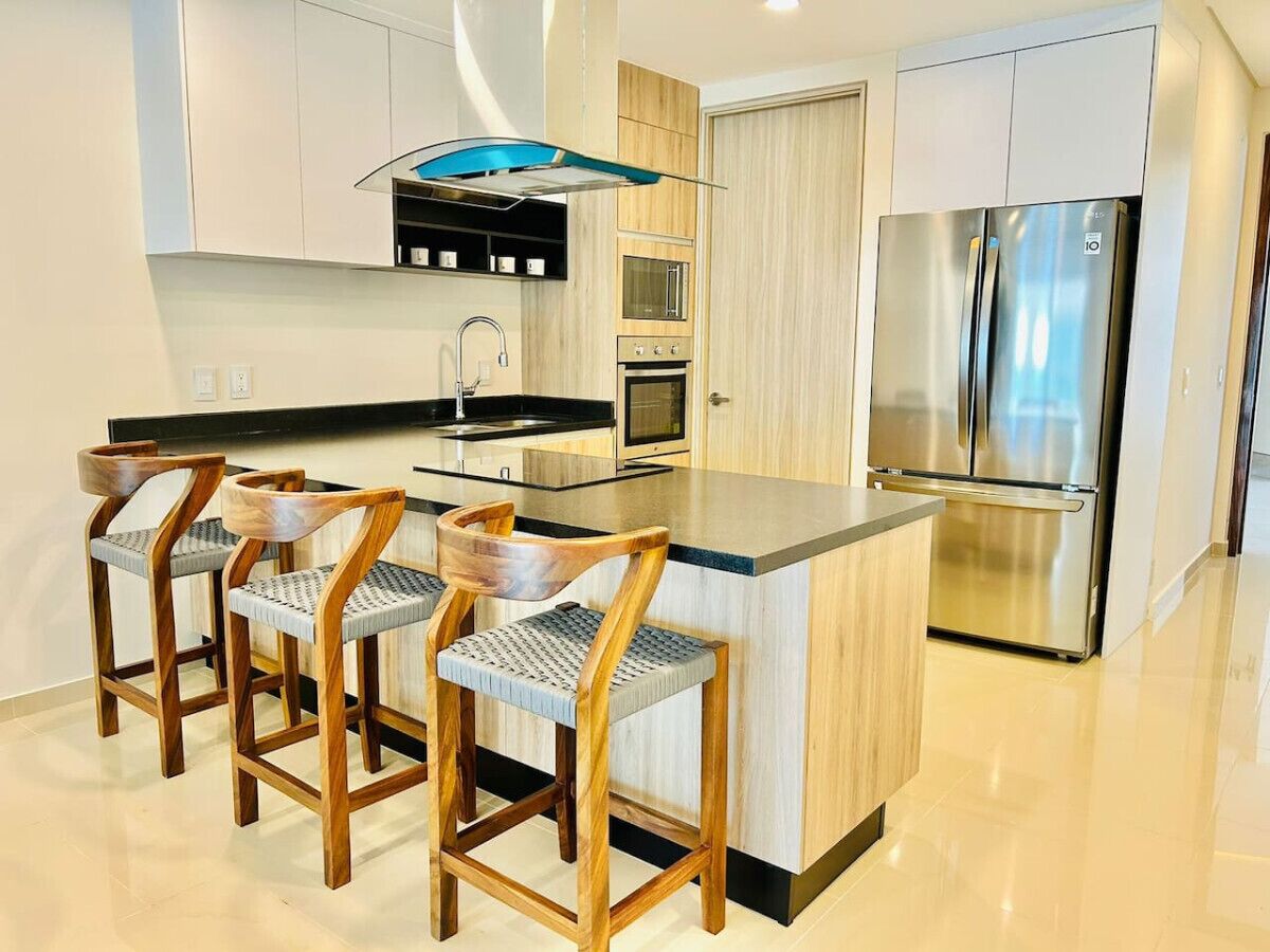 A modern kitchen with a central island, bar stools, stainless steel appliances, and wooden cabinetry.