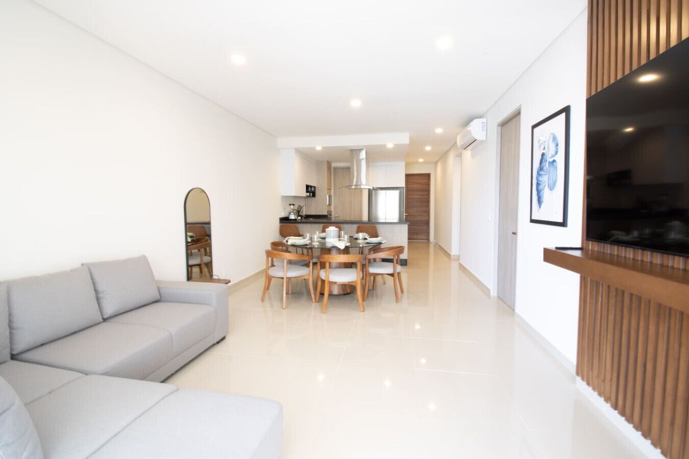 A modern living room with a grey sofa, a dining area with a wooden table and chairs, and a kitchen in the background.