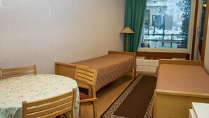 A small, cozy room with two single beds, a wooden table with a floral tablecloth, a chair, a lamp, and a view of the outside through a window. - Kylpylä-hotelli, huone 242 by Interhome (Ikaalinen)