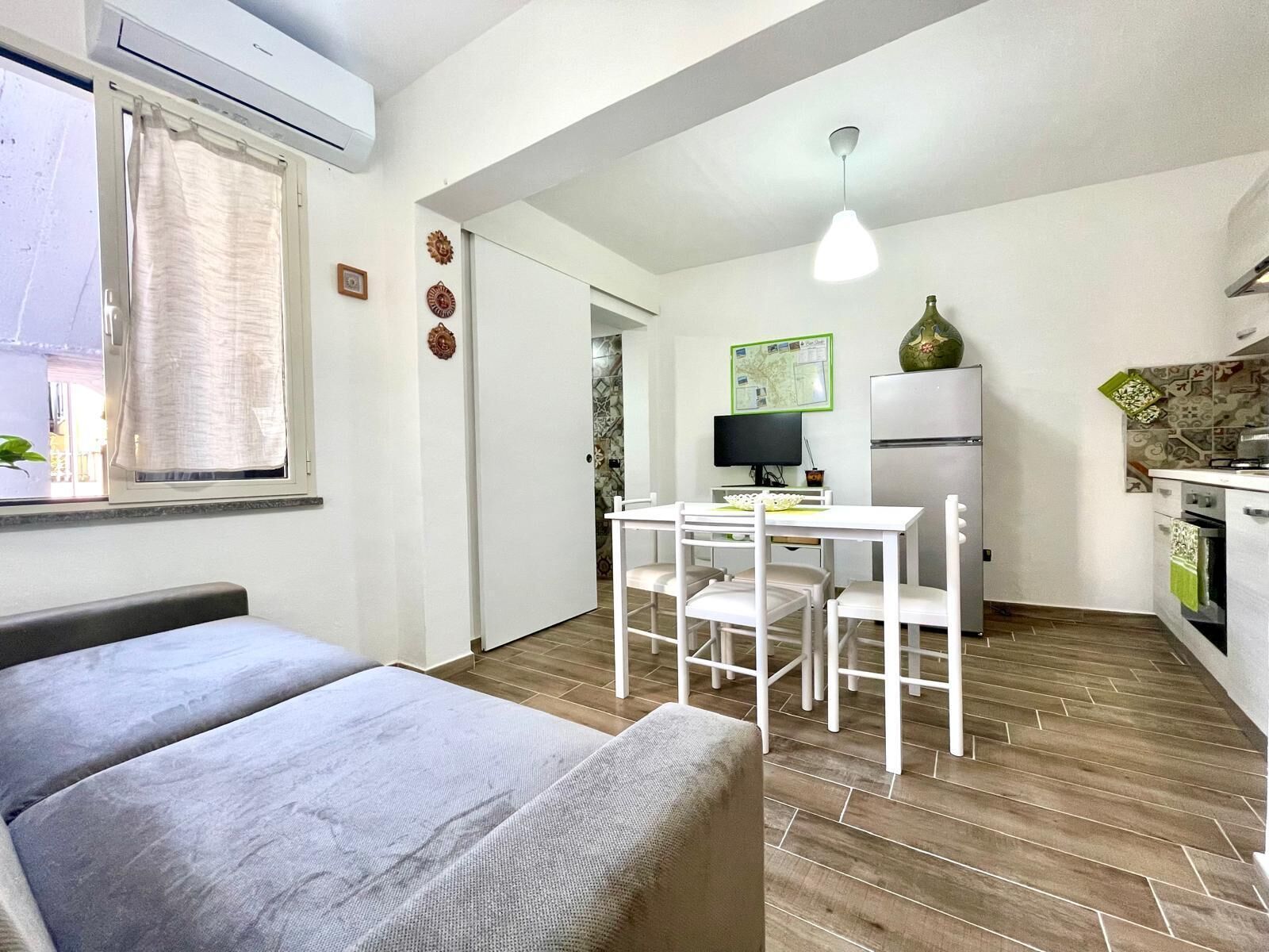 A compact room with a bed, a small dining table with chairs, a kitchen area with a refrigerator, and a window with curtains.