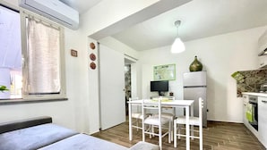 Une pièce de taille modeste comprenant un lit, une petite table à manger avec des chaises, un coin cuisine avec un réfrigérateur et une fenêtre avec des rideaux.