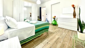 Une chambre d’hôtel comprenant un lit, un bureau, une chaise, un miroir, un vase avec des fleurs et une plante en pot.
