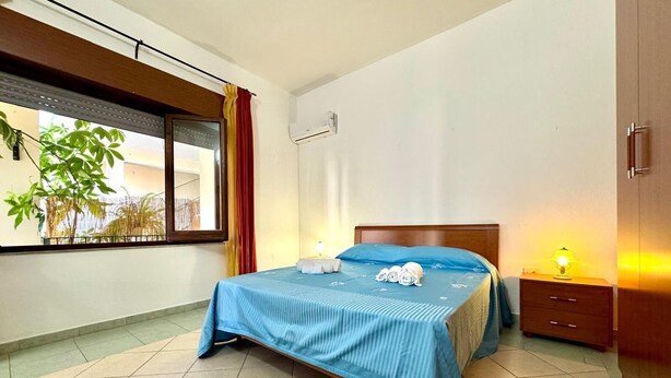 A bedroom with a bed, a nightstand, a lamp, a window with curtains, and a plant. - " La Corte della Pavoncella " (Bari Sardo)