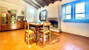 A dining area with a wooden table and chairs, a cabinet with glass doors, a fireplace, and a window with blue shutters. - " La Pergola Deluxe " (Jerzu)